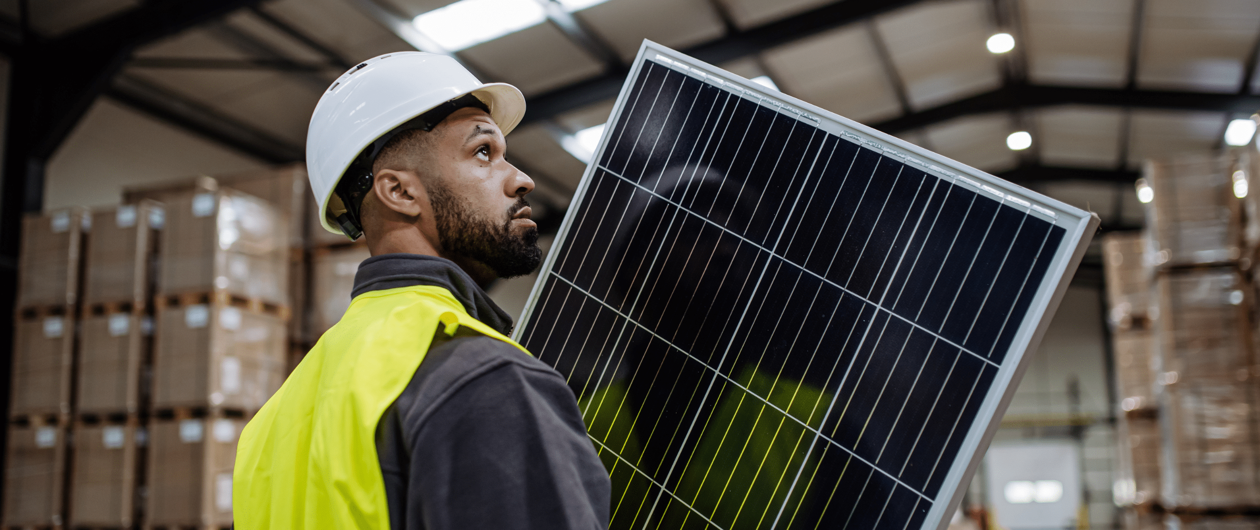 A commercial solar technician in a hard hat and high-visibility vest carefully carries a large solar panel inside an industrial facility, highlighting the scale and professionalism of solar panel recycling operations.