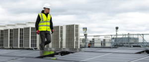 A solar technician wearing a high-visibility vest and hard hat inspects solar panels on a rooftop, with industrial HVAC units and a cloudy sky in the background, illustrating professional solar panel maintenance and recycling operations.