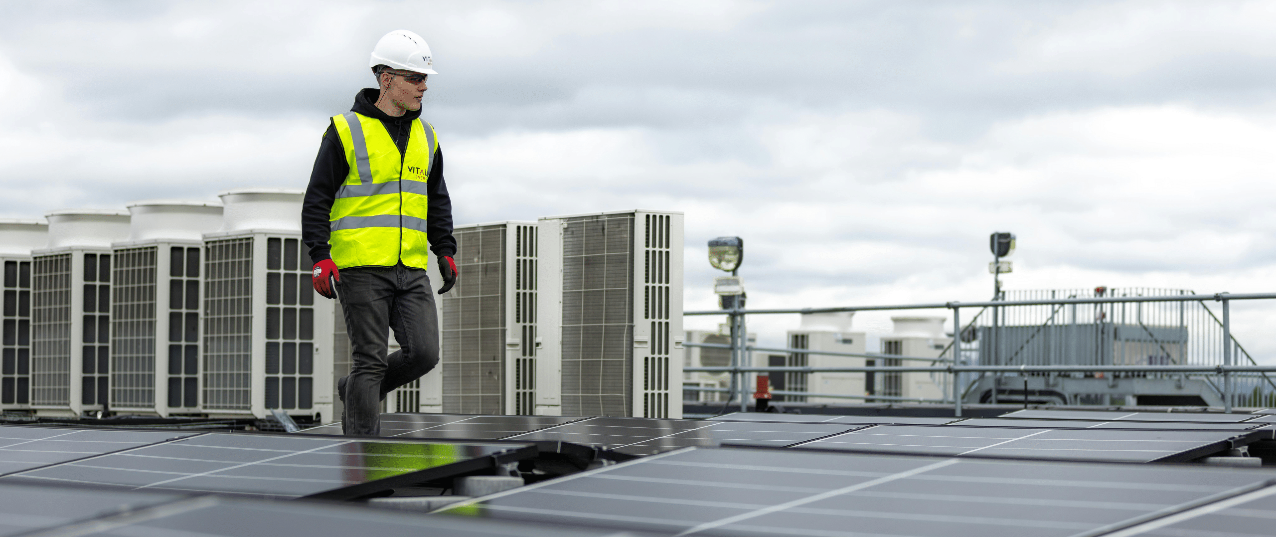 A solar technician wearing a high-visibility vest and hard hat inspects solar panels on a rooftop, with industrial HVAC units and a cloudy sky in the background, illustrating professional solar panel maintenance and recycling operations.