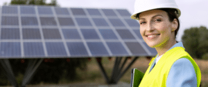 Close-up of a female solar engineer in a hard hat and safety vest, smiling confidently in front of a large solar panel installation, representing solar asset management and sustainability.