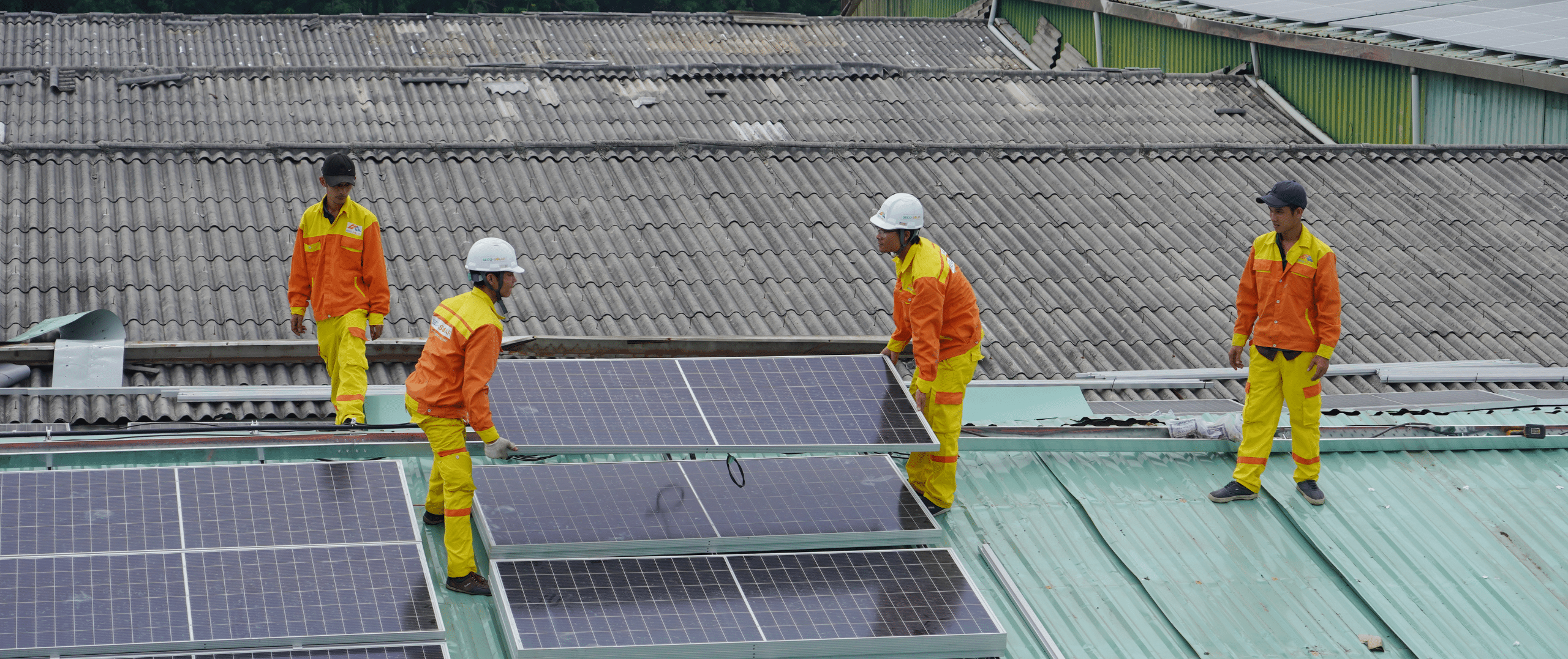 A team of workers in orange and yellow uniforms and safety helmets carefully remove solar panels from a commercial rooftop, illustrating the professional and responsible recycling process for end-of-life solar modules.